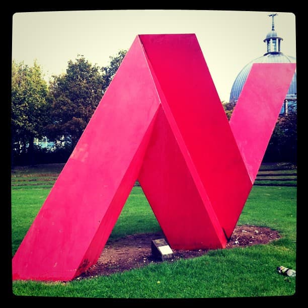 Red painted steel zigzag sculpture on a lawn, with a church dome behind the trees
