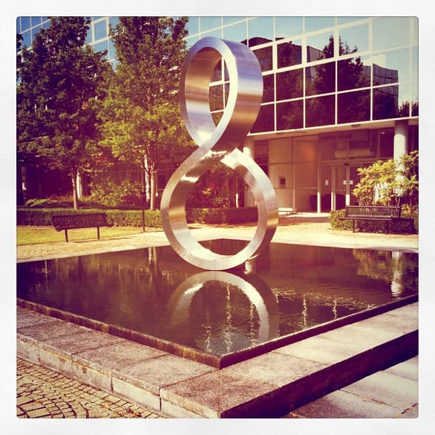 Polished steel figure of eight standing in a square reflecting pool in front of an office building