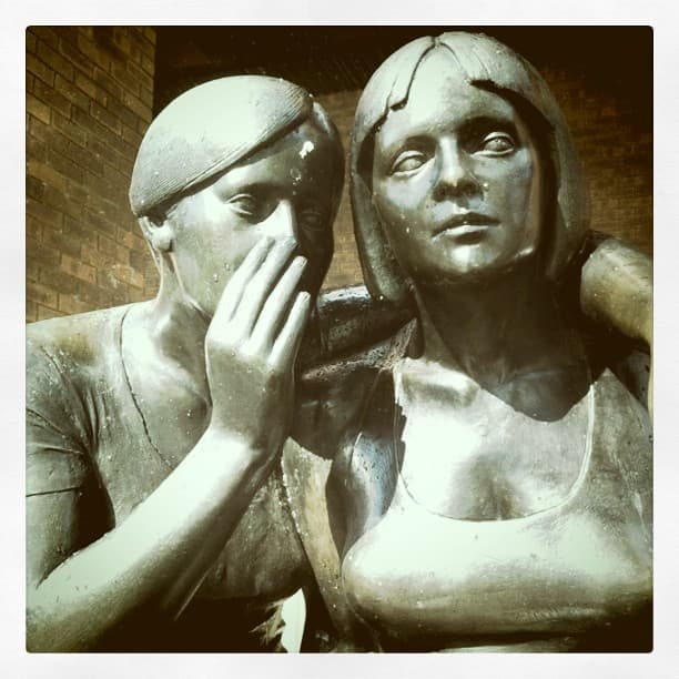 Two bronze figures sitting close, one cupping a hand to whisper in the other's ear, against a brick wall