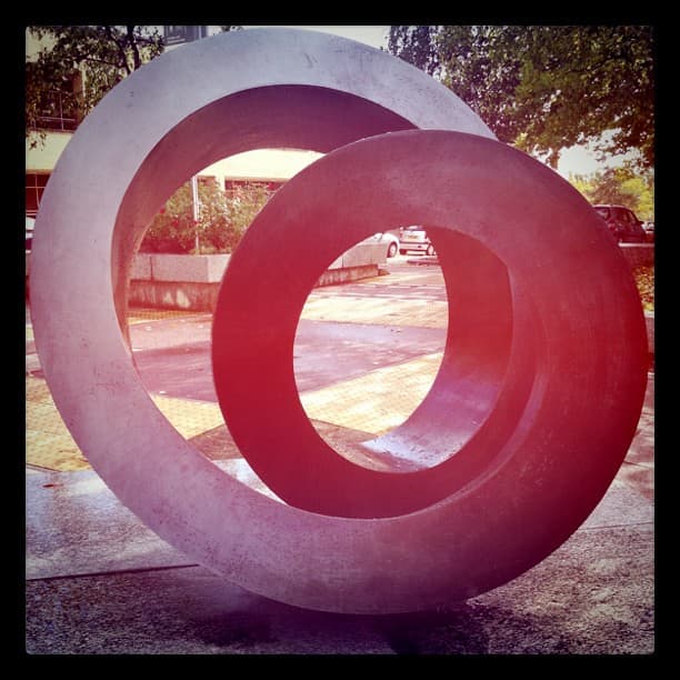 Two large interlocking rings on a paved area, with trees and parked cars behind