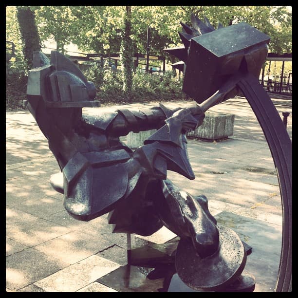 Angular dark bronze figure in a helmet swinging a hammer into a spiked box, in front of a large ring, on a paved square with trees behind