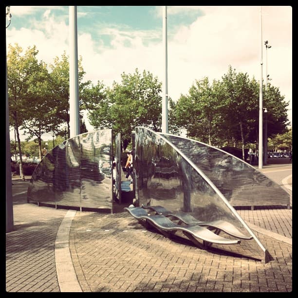 Curved, mirror-polished steel screens and a wavy steel bench on a brick-paved plaza with flagpoles
