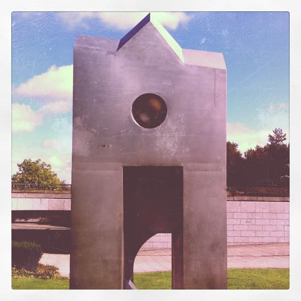 Stainless steel sculpture shaped like a small building with a pitched top, a round window and a doorway, on a lawn