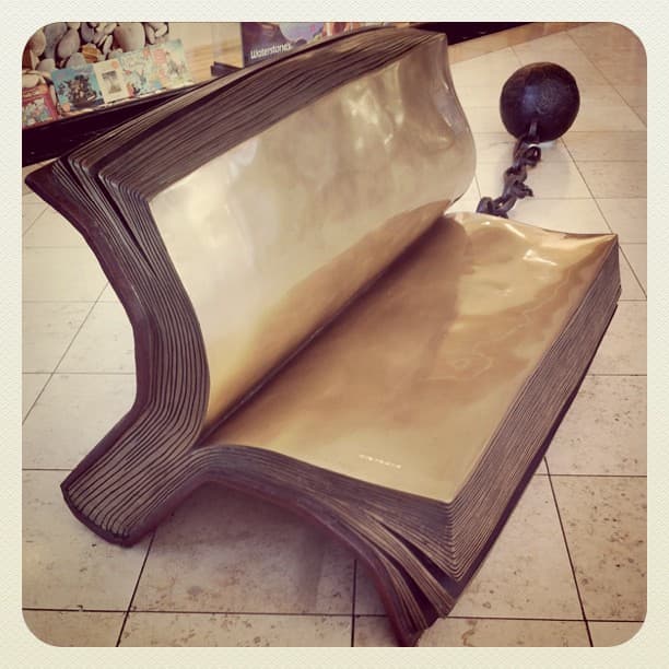 Bronze bench shaped like an open book, chained to a ball, on a tiled shopping-centre floor
