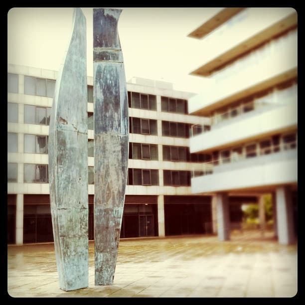 Two tall, slim bronze forms with a green patina in an empty paved square, office buildings behind