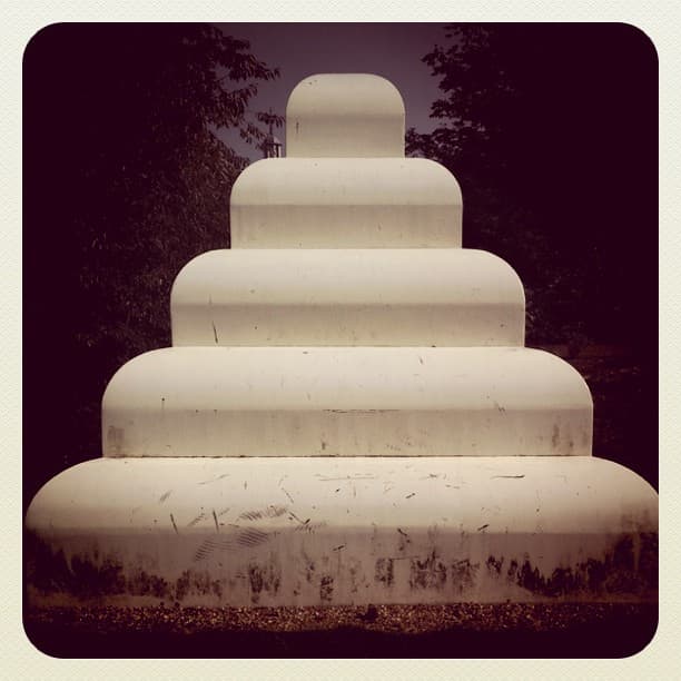 White stepped sculpture of five rounded tiers, scuffed at the base, against dark trees